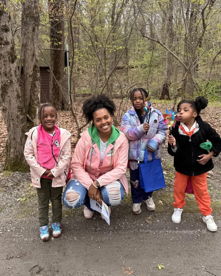 A troop leader kneels beside three young Pearls of Nia Legacy girls on a wooded trail